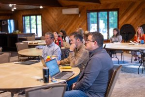 Meeting attendees seated at tables with laptops in a conference room, engaged in discussion and note-taking.