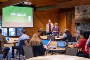 Presenters at West Michigan Trails meeting in a rustic room, speaking to attendees with laptops and notes.