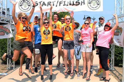 Group of excited runners jumping at a start/finish line during a sunny outdoor event.
