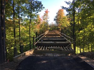 Wooden bridge under construction in a forest with autumn trees around, showcasing a scenic and serene natural environment.