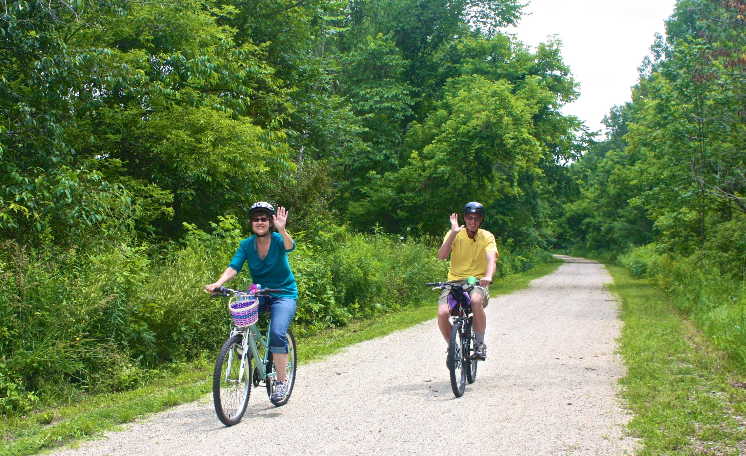 Two people biking on a forest trail, smiling and waving, with lush green trees surrounding them.