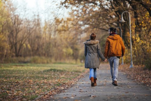 Couple holding hands walking in autumn park, wearing jackets, enjoying a peaceful stroll on a leaf-covered path.