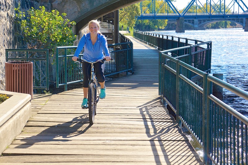 Woman cycling on riverside boardwalk with blue bridge in background on sunny day.