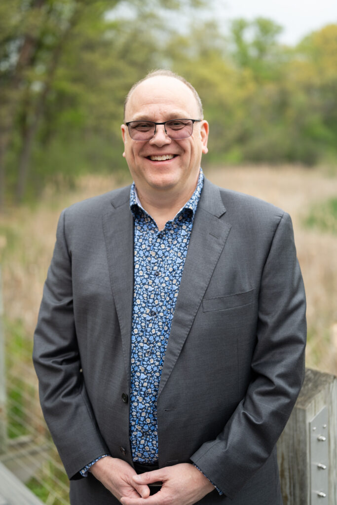 Smiling man in a suit with a floral shirt standing outside on a sunny day.
