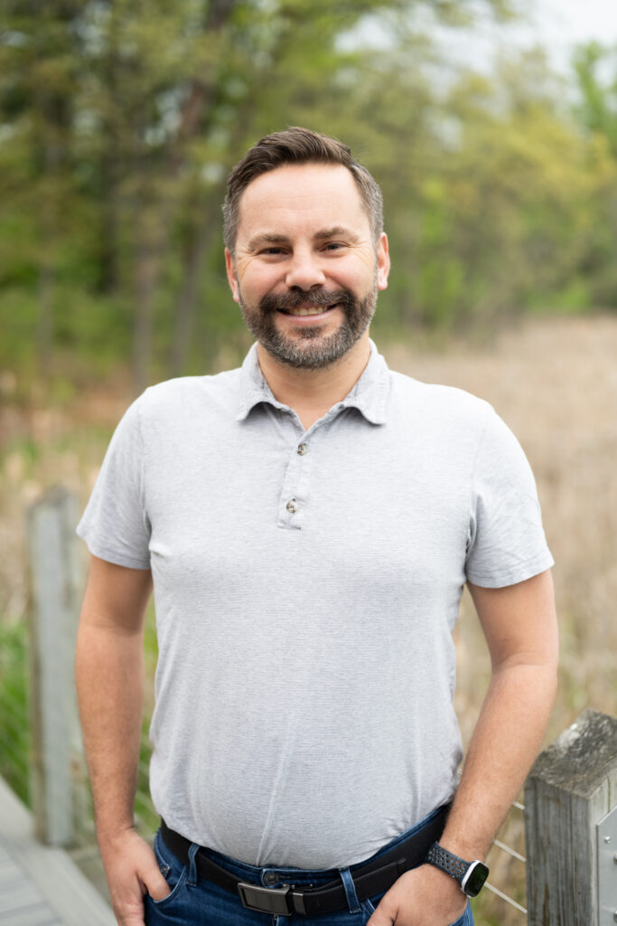 Smiling man in a gray shirt outdoors, standing on a wooden path surrounded by nature.