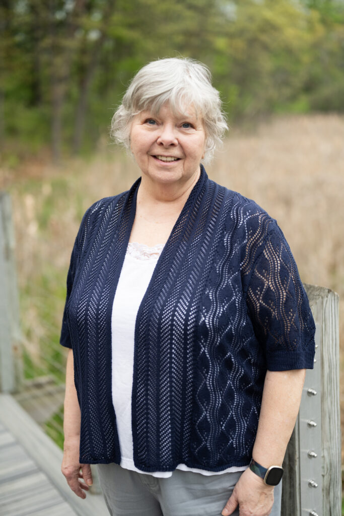 Elderly woman smiling outdoors in a navy cardigan, standing on a boardwalk with trees in the background.