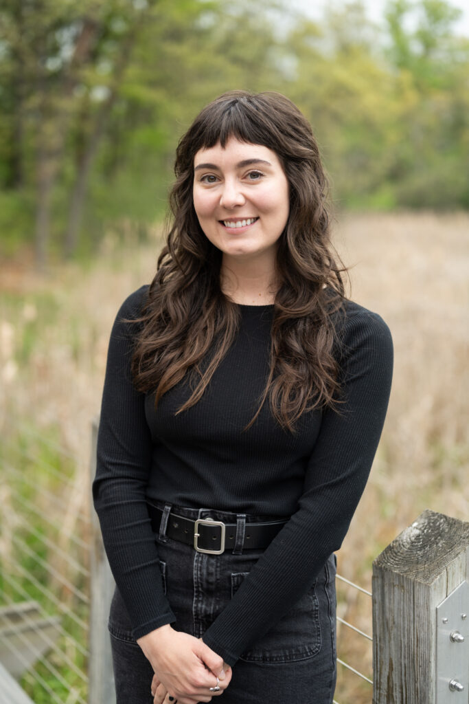 Smiling person in black outfit standing on a wooden walkway with a natural background of trees and grass.