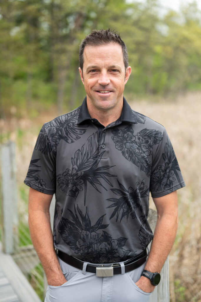 A man in a floral polo shirt, posing outdoors with greenery in the background.