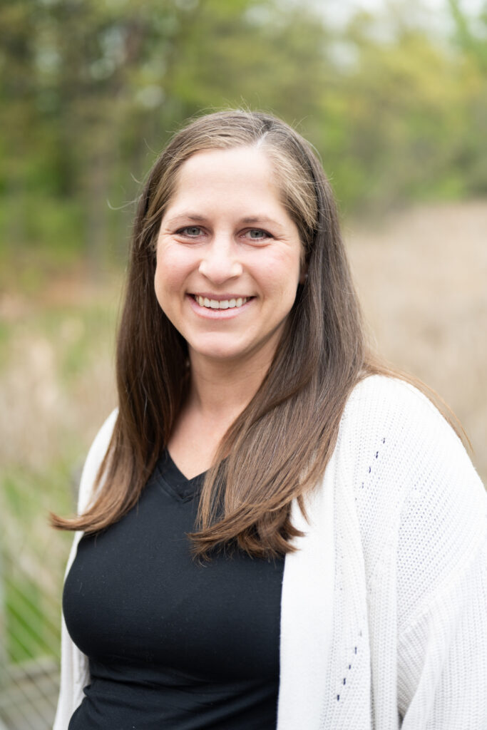 Smiling woman with long hair outdoors wearing a black shirt and white cardigan, green background.