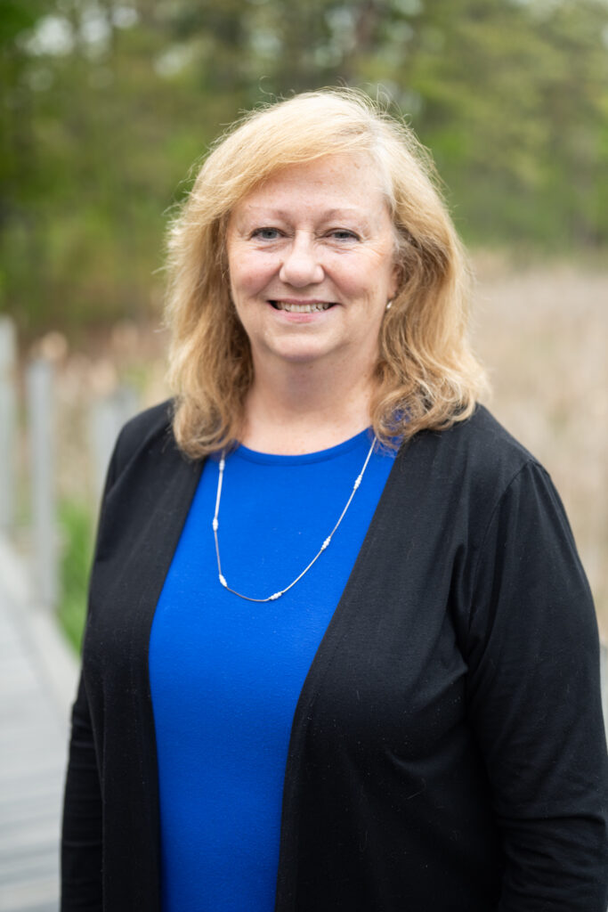 Smiling woman in a blue top and black cardigan standing outdoors with blurred greenery in the background.