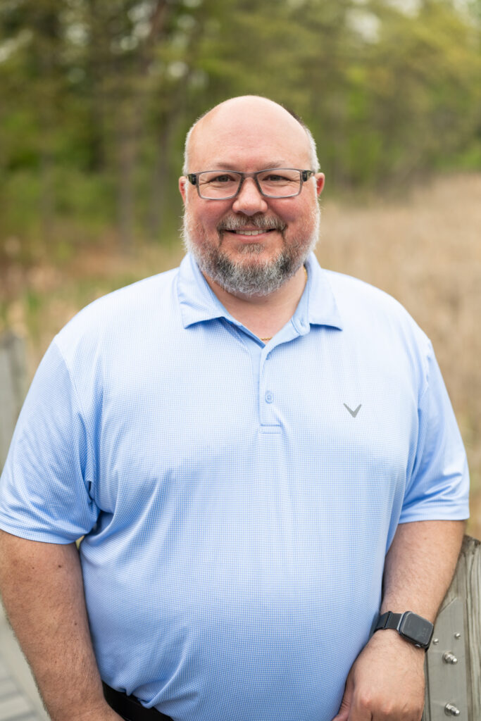 Smiling man in a light blue shirt outdoors, standing confidently with trees in the background.
