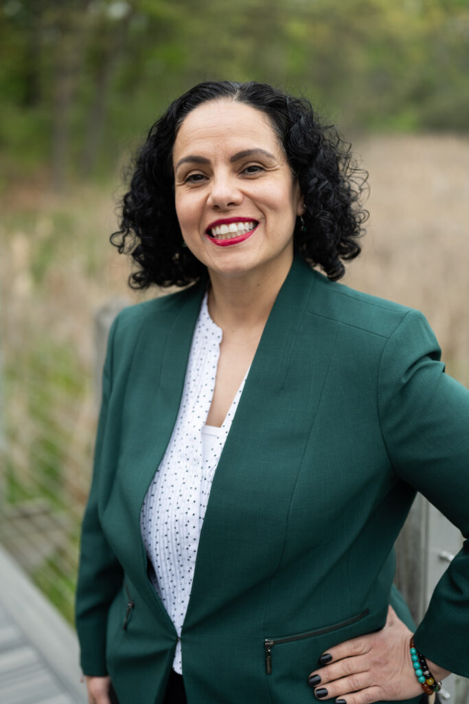 Woman in green blazer smiling outdoors, standing confidently in a natural setting.