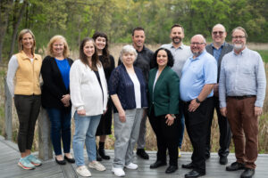 A diverse group of 11 people smiling outdoors on a wooden path surrounded by greenery.