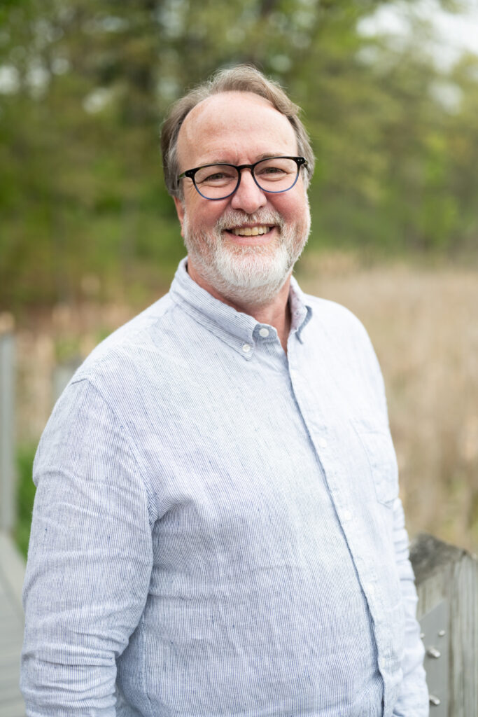 Smiling man with glasses in a light blue shirt, outdoors with blurry green foliage background.