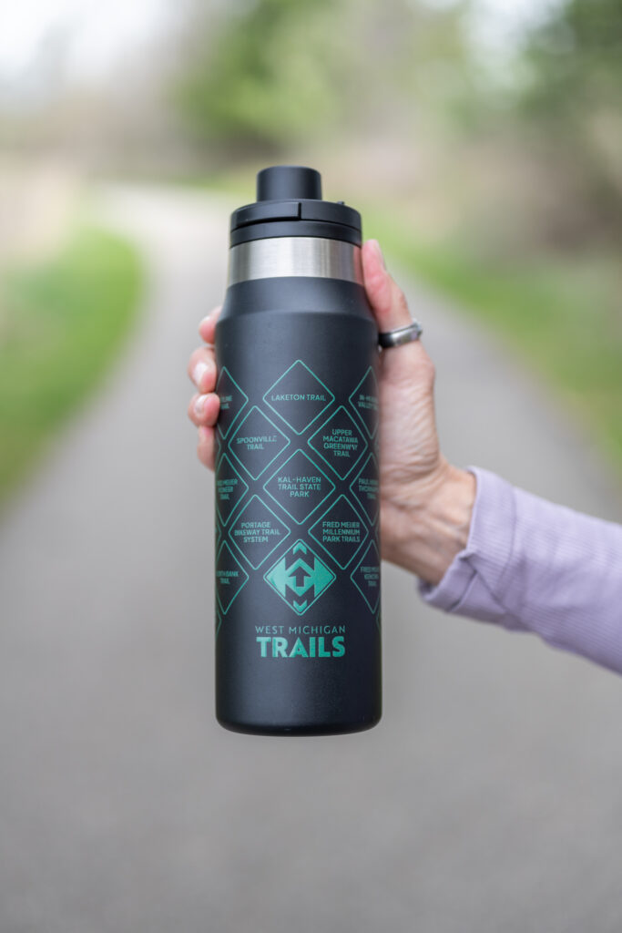 Hand holding West Michigan Trails water bottle on a scenic path, promoting outdoor activities and hydration.