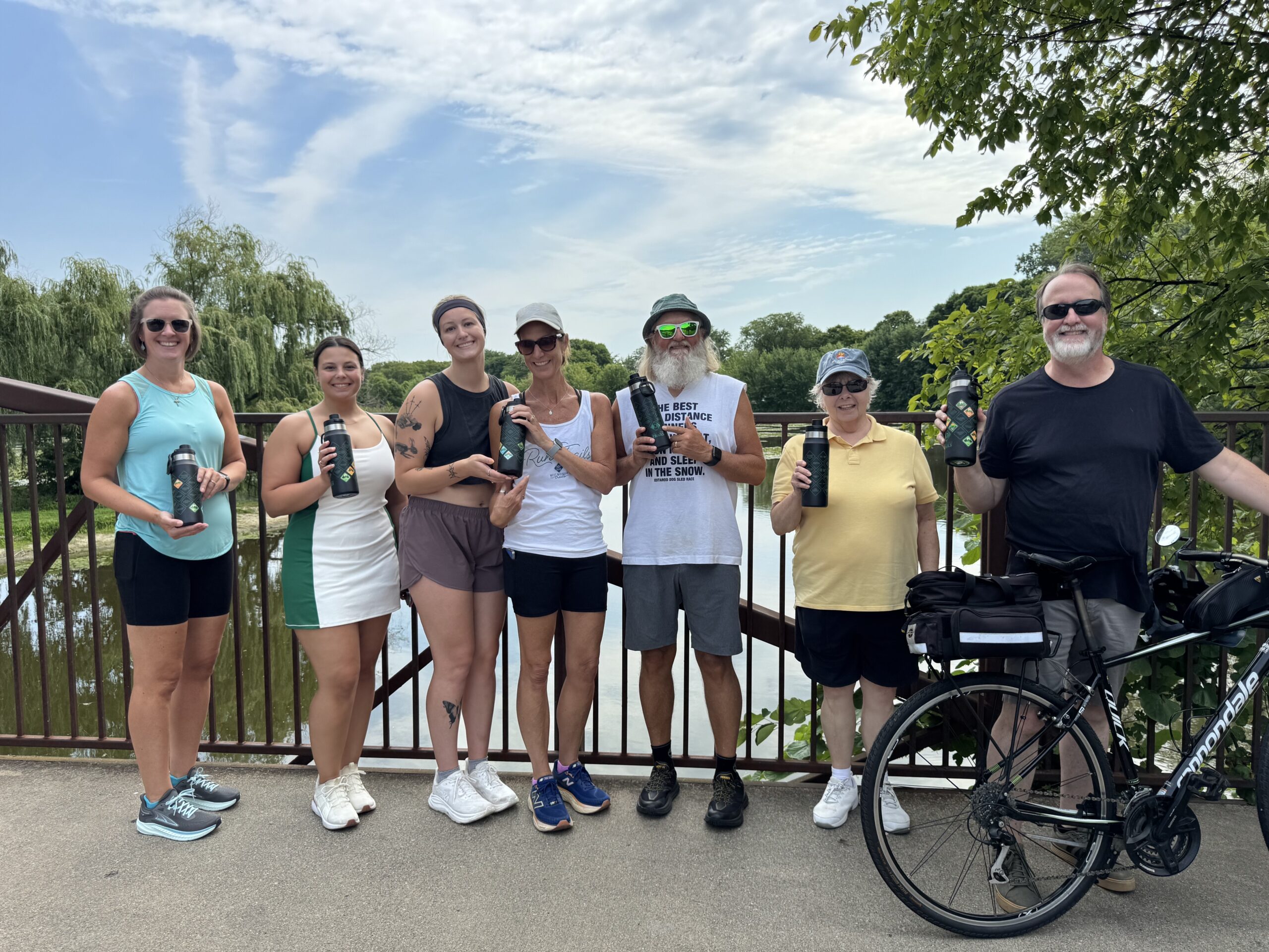 Group on bridge holding water bottles, enjoying outdoor activity with green scenery in background.