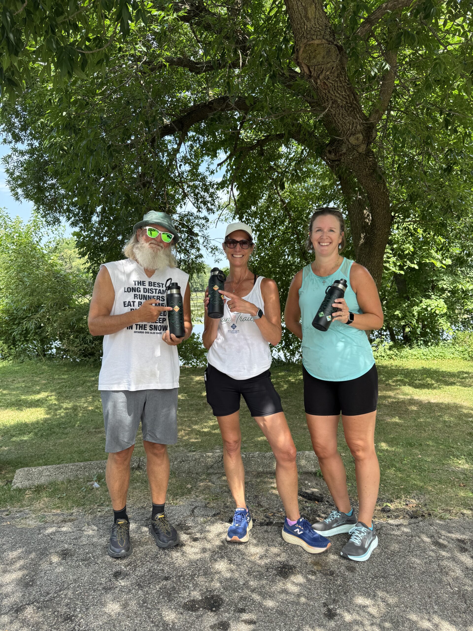 Three people in athletic wear holding water bottles, standing under a tree in a park.