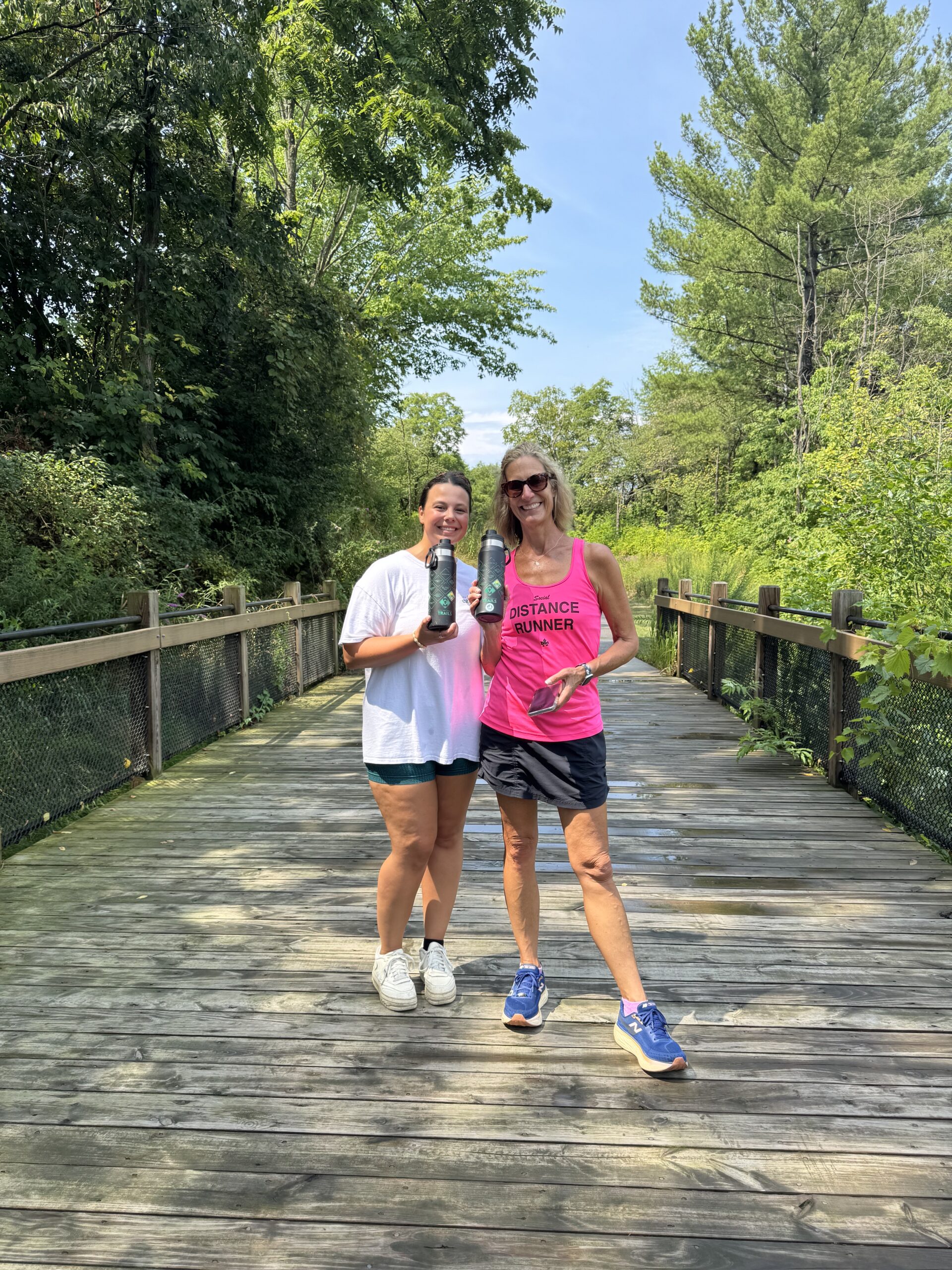 Two women smiling with water bottles on a wooden path surrounded by lush greenery, enjoying a sunny day outdoors.