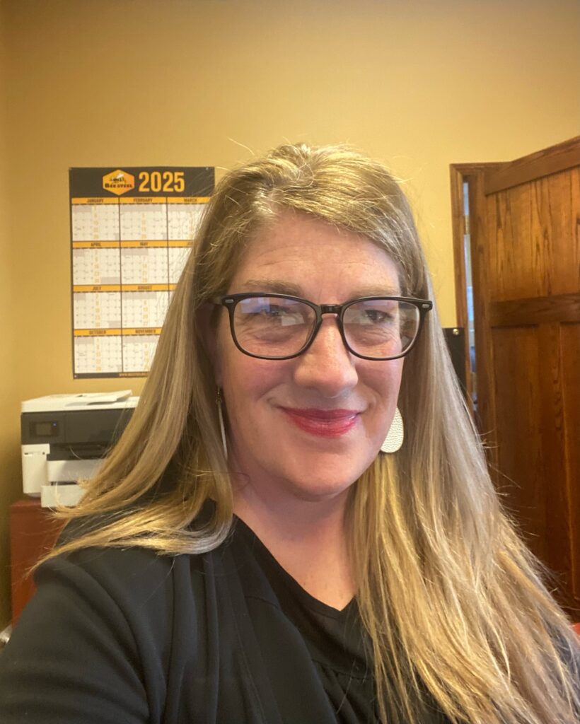 Smiling woman with glasses in office setting, calendar on wall.