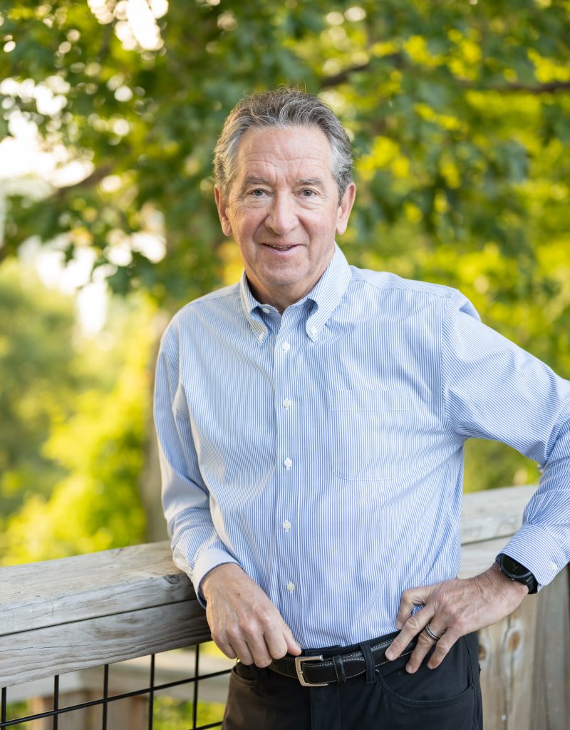 Man in blue shirt smiling outdoors by a wooden fence, surrounded by green trees.