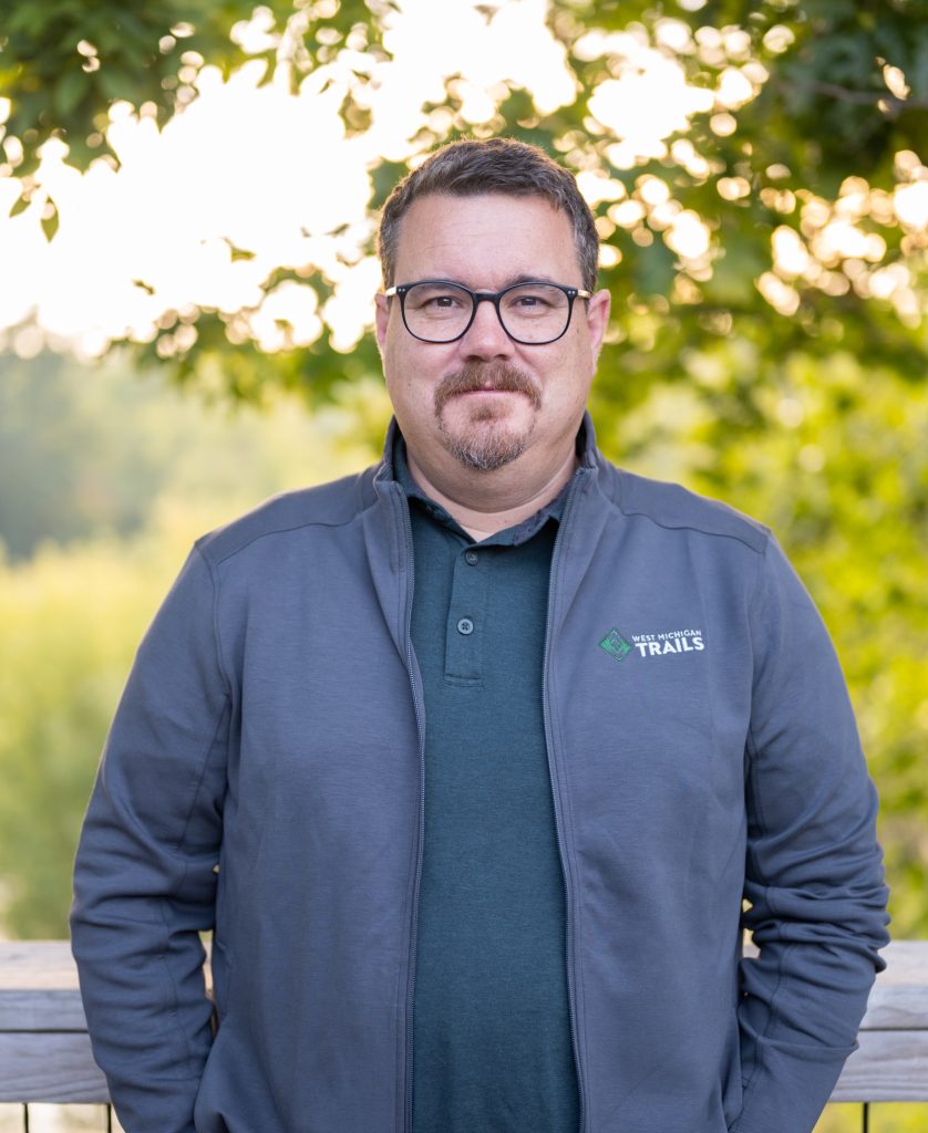 Man in gray jacket and glasses outdoors with blurred green foliage background.