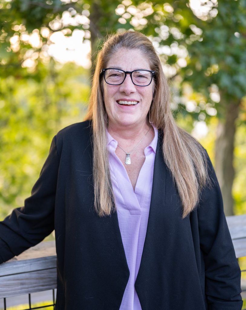 Smiling woman with glasses and long hair outdoors in a black jacket and lavender shirt.