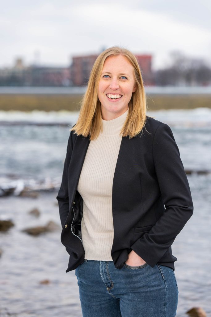 Smiling person in a black jacket by a waterfront on a cloudy day.