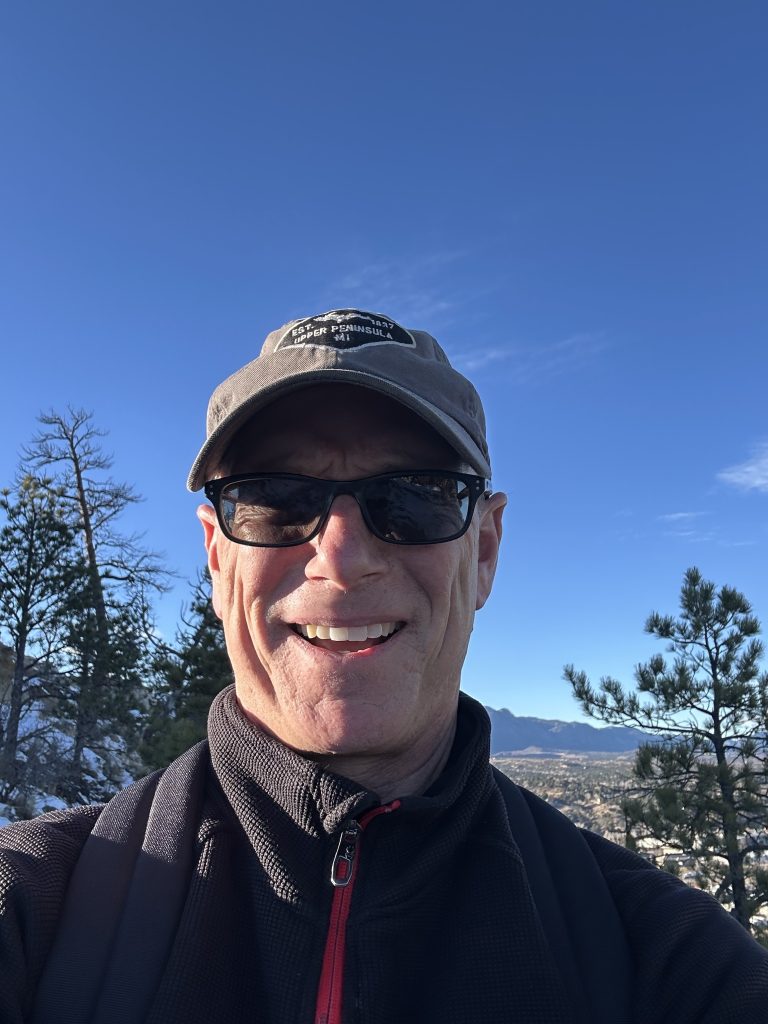 Man enjoying a sunny mountain hike, wearing sunglasses and a baseball cap, with blue sky and trees in the background.