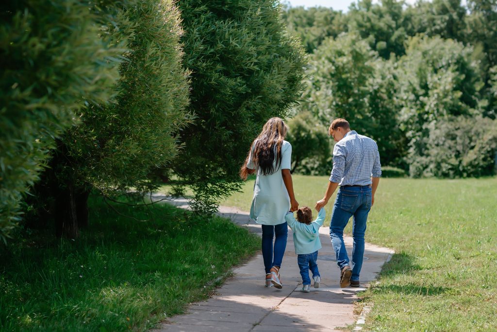 Family walking hand in hand on a sunny park path, surrounded by lush green trees and grass.