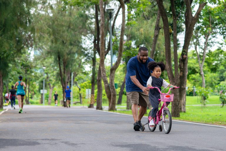 Father teaching daughter to ride a bike with training wheels in a park.