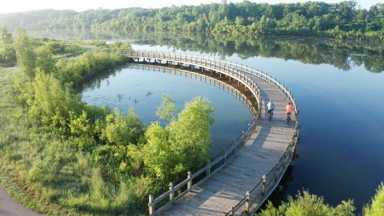 Curved wooden boardwalk over tranquil lake with cyclists, surrounded by lush greenery and scenic landscape.