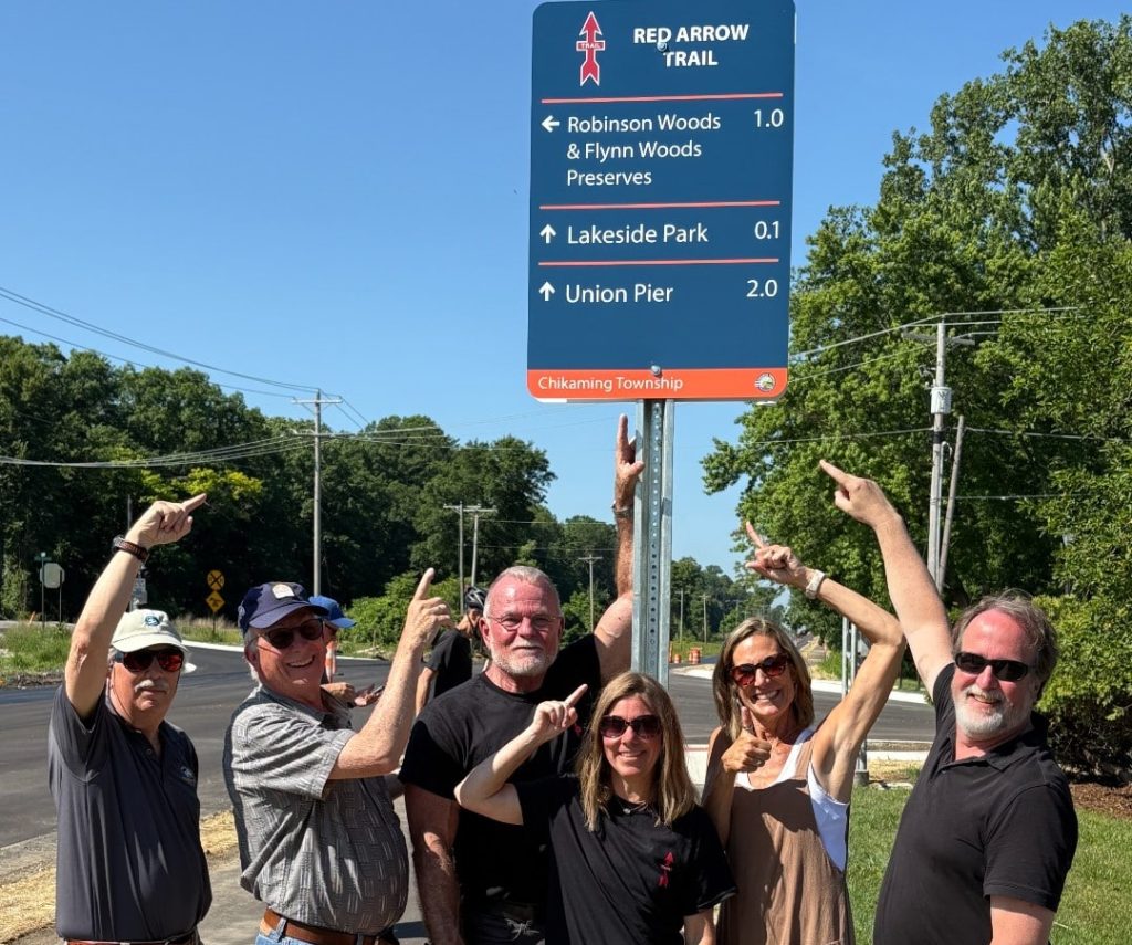 Group of people pointing at Red Arrow Trail sign in Chikaming Township under a clear blue sky.