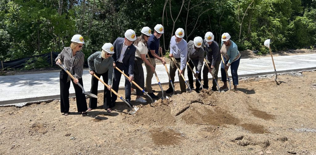 People at a groundbreaking ceremony, wearing hard hats and using shovels, starting construction.