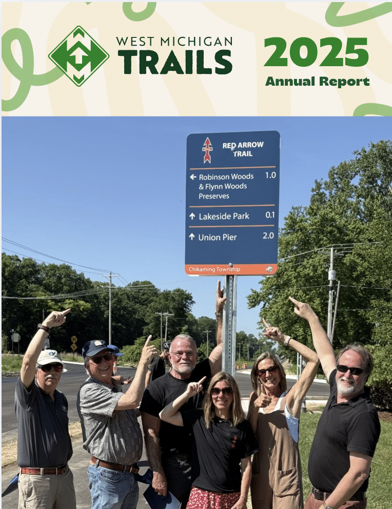 Group points at Red Arrow Trail sign in West Michigan, bright day, trees and road in background.