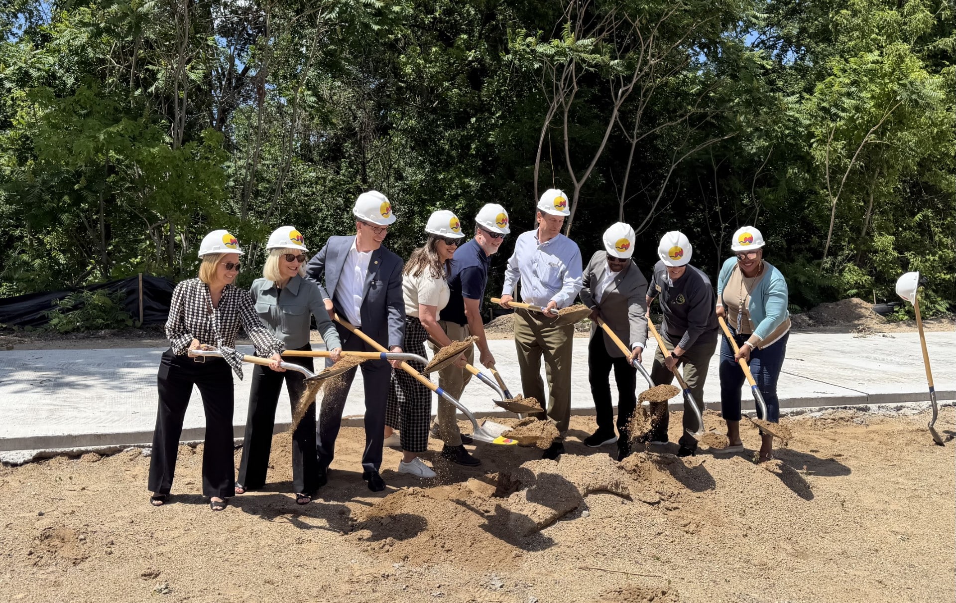 Group of people breaking ground with shovels at a construction site, wearing hard hats and smiling.