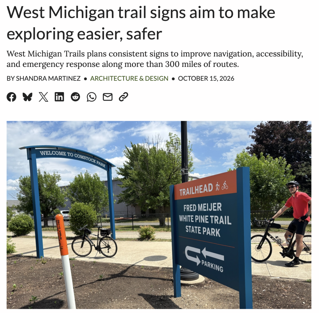 Cyclists at Comstock Park trailhead with signs for Fred Meijer White Pine Trail State Park under a clear blue sky.